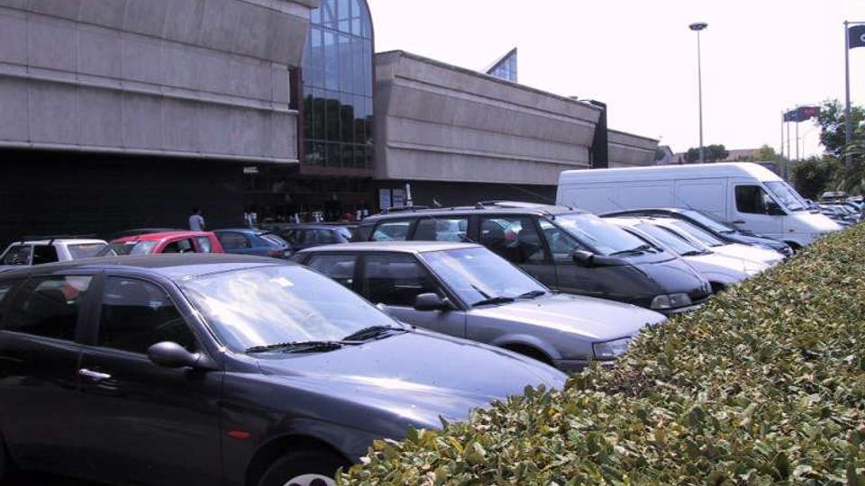 Parcheggio all’aperto affollato di auto e un furgone bianco davanti a un grande edificio moderno in cemento, con una facciata vetrata centrale ad arco. In primo piano si vede una siepe bassa che delimita l’area di sosta.