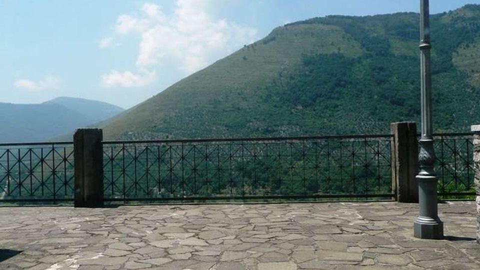 Belvedere pavimentato in pietra con ringhiera in ferro e un lampione, affacciato su una valle boscosa e colline verdi sotto un cielo parzialmente nuvoloso.