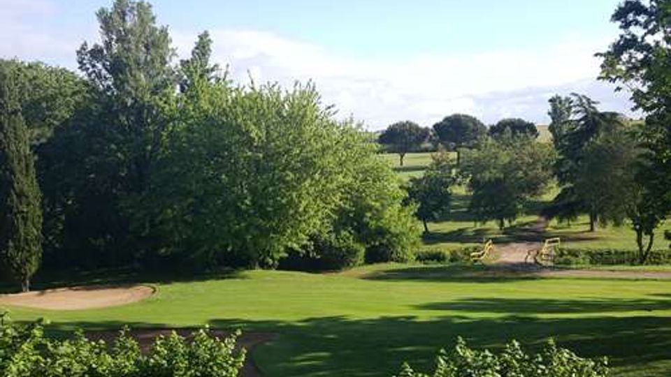 Ampio campo da golf con prato rasato e una buca in primo piano, circondato da alberi e cespugli. In lontananza si vedono ulteriori fairway e zone verdi sotto un cielo sereno.