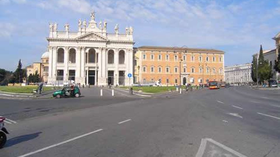 Veduta di una grande piazza urbana con ampia carreggiata e spartitraffico, dominata da una facciata monumentale in stile neoclassico con colonne, arcate e statue sul coronamento. Sullo sfondo si vedono edifici storici allineati lungo la strada, con cielo parzialmente nuvoloso.