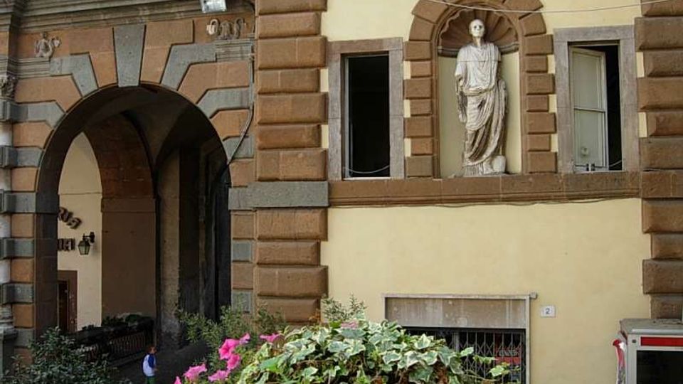 Facciata di un edificio storico con grandi blocchi bugnati in pietra e un ampio arco d’ingresso che conduce a un passaggio coperto. Al piano superiore, una statua in una nicchia arcuata è affiancata da finestre incorniciate in pietra, con vegetazione e fiori in primo piano.