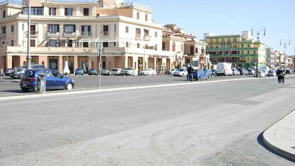 Ampio viale urbano con carreggiata larga e auto parcheggiate lungo il bordo, fiancheggiato da edifici residenziali a più piani con facciate chiare, balconi e portici al piano terra. Sullo sfondo si vedono palazzi colorati e lampioni stradali, sotto un cielo sereno.