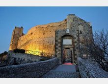 Veduta di un castello medievale in pietra su un’altura, con alte mura illuminate al crepuscolo. In primo piano si vede l’ingresso ad arco con portale e una passerella in pietra delimitata da muretti e ringhiere.