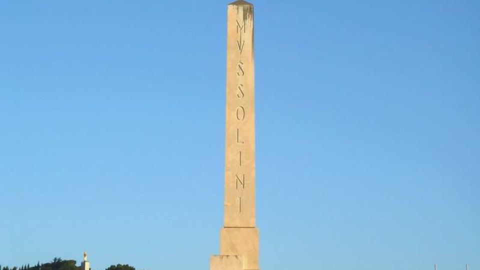 Obelisco in pietra chiara con incisioni verticali, collocato al centro di una piazza aperta. Sullo sfondo si vedono un grande stadio con struttura bianca e alberi, sotto un cielo sereno.