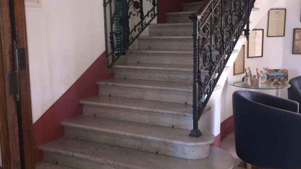 Scala interna in marmo con ampi gradini curvi e ringhiere in ferro battuto decorato, all’interno di un edificio con pareti bianche e fascia bordeaux. Sulle pareti sono appesi quadri e cornici, con una piccola area salotto e pavimento in marmo policromo.