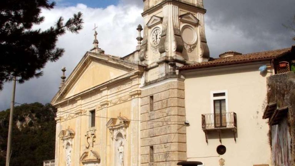 Chiesa in stile barocco con facciata color ocra e campanile in pietra sormontato da cupola, affacciata su una piccola piazza con una fontana centrale. Sullo sfondo si vedono edifici adiacenti e alberi, sotto un cielo nuvoloso.
