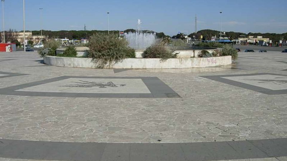 Ampia piazza pavimentata in pietra con motivi geometrici, al centro una fontana circolare rialzata con getti d’acqua e aiuole di vegetazione. Sullo sfondo si vedono lampioni alti, strade e costruzioni basse in un’area aperta.