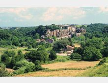 Veduta panoramica di un borgo con edificio fortificato e mura in pietra arroccati su uno sperone roccioso, circondati da fitte aree boschive. In primo piano si vedono campi coltivati e alcune case rurali sparse nella valle.
