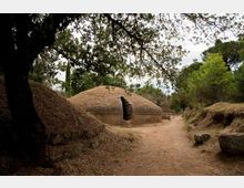 Sentiero sterrato in un’area boschiva che passa tra tumuli erbosi e strutture in pietra a pianta circolare con copertura a cupola, parzialmente interrate. Alberi e vegetazione circondano il percorso, con cielo nuvoloso sullo sfondo.