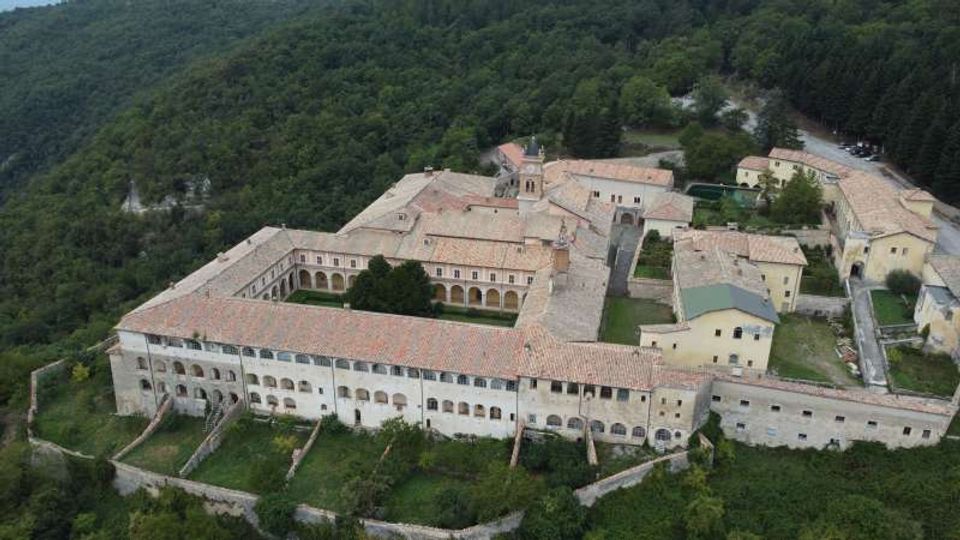Veduta aerea di un grande complesso monastico in pietra con tetti in coppi rossi e chiostro interno, disposto attorno a cortili e passaggi ad arcate. L’edificio è circondato da un muro di cinta e immerso in un paesaggio collinare boscoso.
