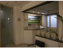 Interno di un ufficio con area reception: bancone in muratura, scaffali con raccoglitori verdi e blu e illuminazione a faretti nel soffitto. A lato si vedono una porta in vetro satinato e una finestra, con un piccolo piano d’appoggio con contenitori trasparenti.