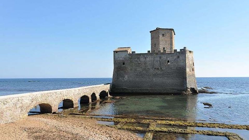 Fortezza in pietra sul mare, collegata alla riva da un ponte rialzato con più arcate. Davanti si vede una spiaggia rocciosa con pozze d’acqua e, sullo sfondo, l’orizzonte del mare sotto un cielo sereno.