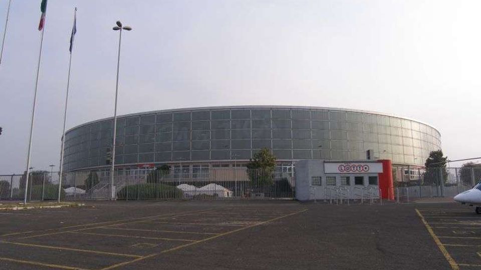 Grande edificio moderno di forma ovale con facciata in vetro e pannelli metallici, recintato e affacciato su un ampio parcheggio asfaltato con segnaletica a terra. In primo piano si vedono un piccolo ingresso con insegna e alcune aste portabandiera e lampioni.