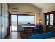 Camera da letto con pavimento in legno, armadio azzurro e letto con copriletto blu, affacciata su una grande porta finestra scorrevole che conduce a un terrazzo con vista panoramica. Nella stanza ci sono una scrivania con due sedie, tende bianche e una finestra laterale con inferriate decorative.