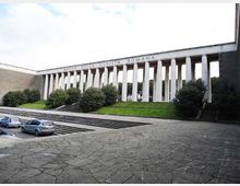 Facciata del Museo della Civiltà Romana con lunga colonnata di pilastri bianchi e scritta in sommità, affacciata su una piazza pavimentata con ampie scalinate. Davanti si vedono aiuole verdi con arbusti e un’area di parcheggio ai margini.