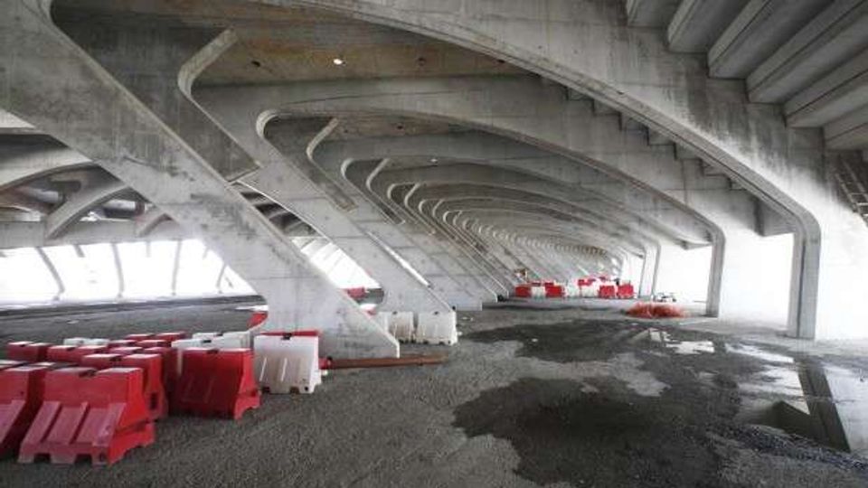 Spazio sotto un grande viadotto in cemento armato, con una serie di archi e pilastri inclinati ripetuti in prospettiva. Sul pavimento in ghiaia e asfalto con pozzanghere sono presenti barriere stradali in plastica rosse e bianche disposte lungo i lati.