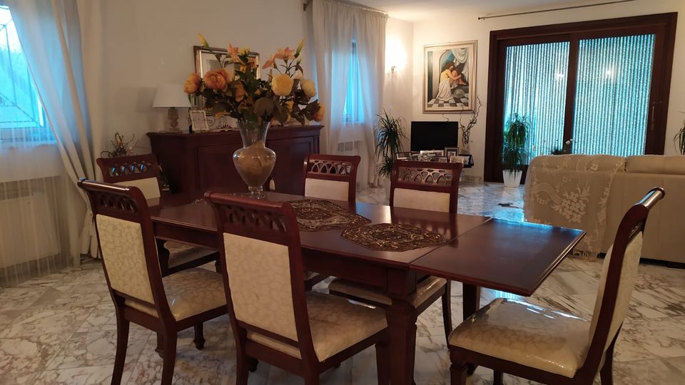 Soggiorno con zona pranzo: grande tavolo in legno scuro con sedie imbottite e centrotavola floreale, su pavimento in marmo chiaro. Sullo sfondo ci sono finestre con tende leggere, una credenza, un quadro, piante e una porta finestra con vetrate rigate accanto a un divano.