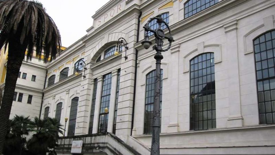 Facciata di un edificio storico con ampie finestre ad arco e cornici in pietra, con scalinata esterna e ingresso sotto un grande arco con scritta “SPQR”. In primo piano si vede un lampione ornamentale e vegetazione con palme, in un contesto urbano.