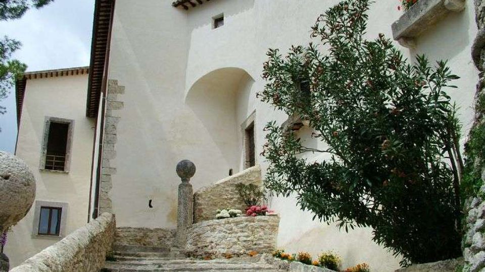 Scalinata in pietra che sale tra edifici intonacati di bianco con tetti in coppi e un arco di passaggio. Ai lati ci sono muretti in pietra, una colonna con sfera decorativa e un albero con aiuole fiorite vicino alle finestre.