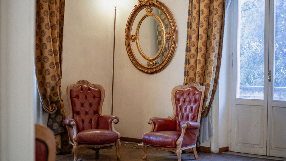 Salotto elegante con due poltrone in pelle rossa, grande tappeto decorato e pavimento in parquet. Lampadario in cristallo e specchio ovale dorato sulla parete, con alte finestre a portafinestra incorniciate da tende pesanti.