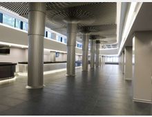 Ampio atrio interno di un edificio moderno con pavimento in grandi piastrelle scure, illuminazione lineare a soffitto e una fila di colonne metalliche scanalate con capitelli a raggiera. Ai lati si vedono banconi bassi e finestre alte che lasciano entrare luce naturale, con vista verso un’area vetrata in fondo.