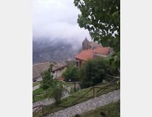 Vista di un borgo collinare con tetti in tegole rosse e una piccola chiesa in pietra con campanile, affacciati su una valle avvolta dalla nebbia. In primo piano si vedono un sentiero in ciottoli, staccionate in legno, vegetazione e rami di alberi che incorniciano la scena.
