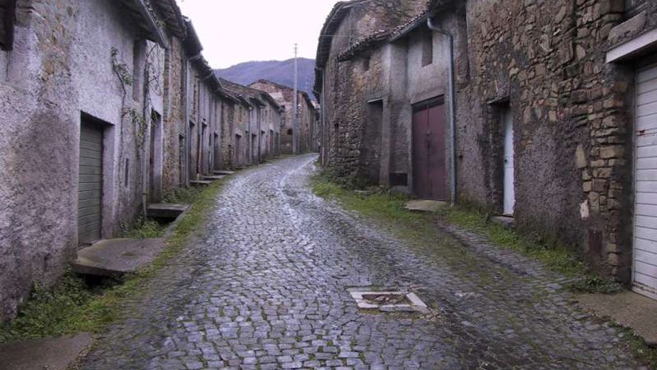 Strada stretta in acciottolato che sale tra case rurali in pietra e intonaco, con portoni e piccole finestre affacciati sulla via. Sullo sfondo si vedono colline e un lampione sospeso sopra la carreggiata.