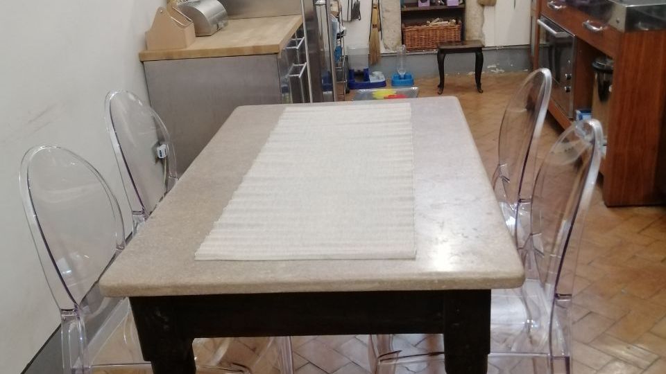 Cucina abitabile con grande tavolo rettangolare e sedie trasparenti, pavimento in cotto a spina di pesce e pareti decorate con quadri. Sul fondo si vede un camino con cappa e rivestimenti di piastrelle colorate, affiancato da elettrodomestici e piano cottura con pensili in legno.