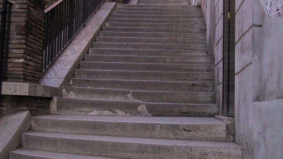 Scalinata esterna in cemento che sale tra edifici in muratura, con ringhiera metallica e muretti in mattoni sul lato sinistro. In cima si intravedono alberi e cielo, mentre a destra corre una parete intonacata con una porta.