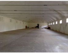 Ampio capannone o magazzino vuoto con pavimento in cemento e soffitto alto attraversato da lunghe travi e cavi. Pareti bianche con una fila di piccole finestre sul lato destro e alcuni materiali accatastati in fondo vicino a una porta.