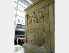 Grande pannello in pietra con bassorilievi figurativi e motivi floreali intrecciati, incorniciato da decorazioni scolpite e fregi a greca. L’opera è esposta in un ambiente museale con parete vetrata e soffitto in vetro che lasciano intravedere l’esterno.