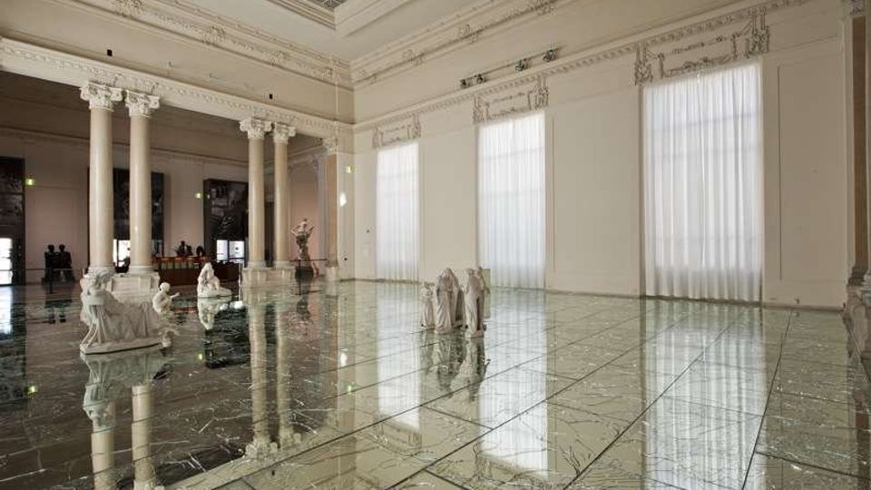 Ampia sala interna in stile neoclassico con colonne e cornici ornamentali, grandi finestre con tende bianche e soffitto alto. Il pavimento lucido a specchio riflette l’architettura e alcune sculture disposte nello spazio.