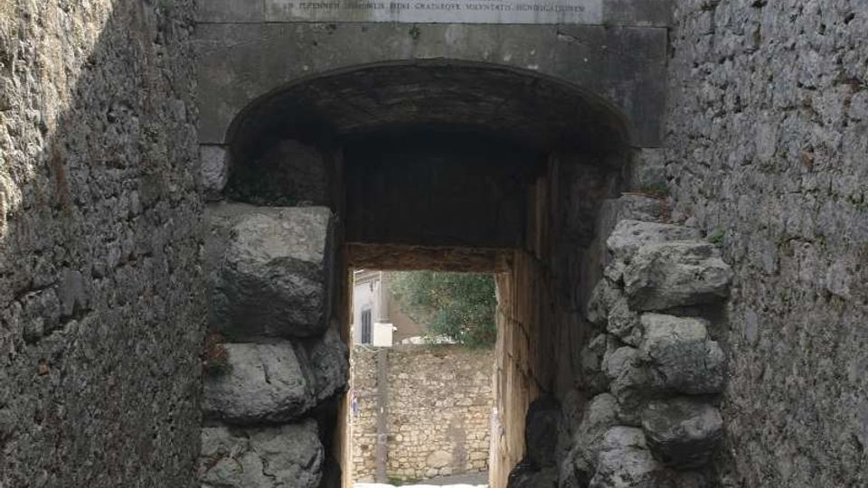 Passaggio in discesa con pavimentazione in ciottoli che attraversa un arco in pietra tra alti muri di mura antiche. Sopra l’arco è visibile una grande epigrafe in marmo con iscrizione, con vegetazione sullo sfondo.