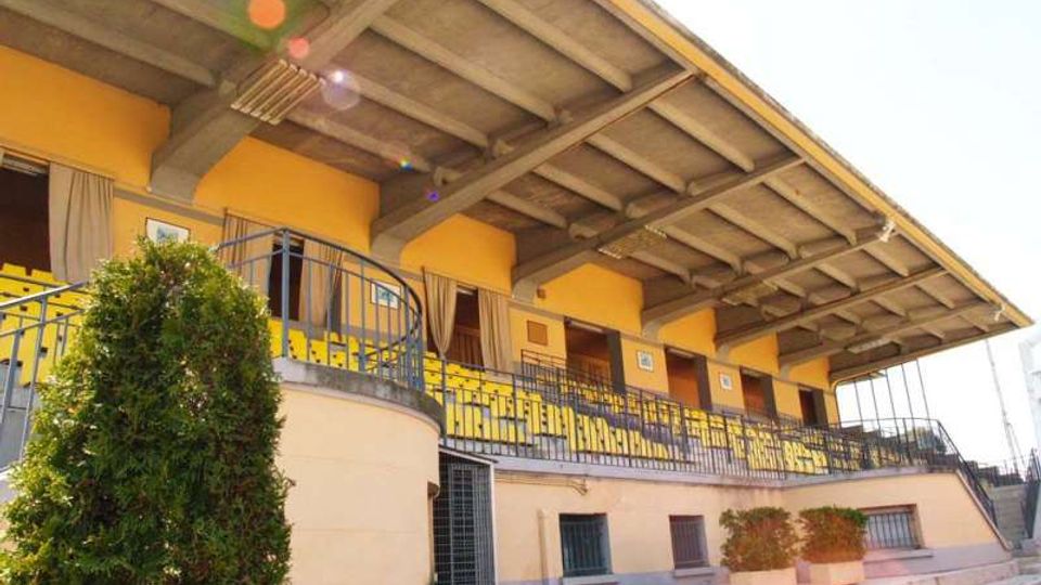 Tribuna coperta di un impianto sportivo con struttura in cemento e tetto sporgente, file di sedute gialle e ringhiere metalliche lungo il camminamento. In primo piano si vedono gradinate in cemento e alcune piante in vaso vicino all’ingresso.