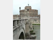 Ponte in pietra con parapetto decorato da statue e lampioni che attraversa un fiume, visto in prospettiva lungo le arcate. Sullo sfondo si innalza una grande fortezza circolare in muratura con edificio superiore e statua sulla sommità.