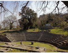 Anfiteatro all’aperto in pietra con gradinate semicircolari su un prato, circondato da alberi e vegetazione su un pendio. Sullo sfondo si vedono un piccolo muro in pietra e il cielo tra i rami spogli.