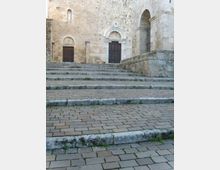 Scalinata in pietra che conduce a una facciata storica in blocchi di pietra chiara, con due portali ad arco e porte in legno scuro. A destra si vede un grande arco d’ingresso con parapetto in pietra e una finestra stretta sopra uno dei portali.