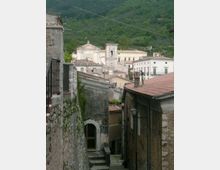 Vicolo in discesa tra edifici in pietra e intonaco con tetti in coppi, che conduce verso il centro di un borgo. Sullo sfondo si vedono una chiesa con facciata classica e un campanile, con colline boscose tutt’intorno.