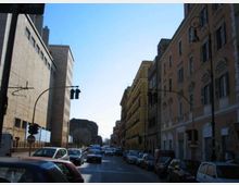 Strada urbana rettilinea con molte auto in sosta e in transito, fiancheggiata da edifici alti: a destra palazzi storici con finestre ad arco, a sinistra un grande edificio moderno. Sono visibili semafori e lampioni lungo la carreggiata sotto un cielo sereno.