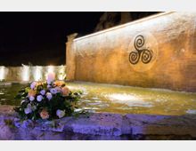 Fontana notturna con vasca d’acqua illuminata e parete in pietra sullo sfondo, decorata da un grande simbolo a spirale. In primo piano sul bordo in pietra c’è un mazzo di fiori con una candela accesa.