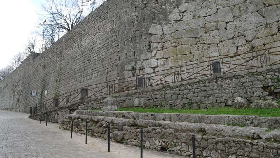Lungo muro di cinta in pietra con tessitura irregolare, affiancato da una strada lastricata e da terrazze in muratura a gradoni con piccoli tratti d’erba. Una rampa laterale con parapetto metallico costeggia il muro, con alberi spogli visibili sopra la sommità.