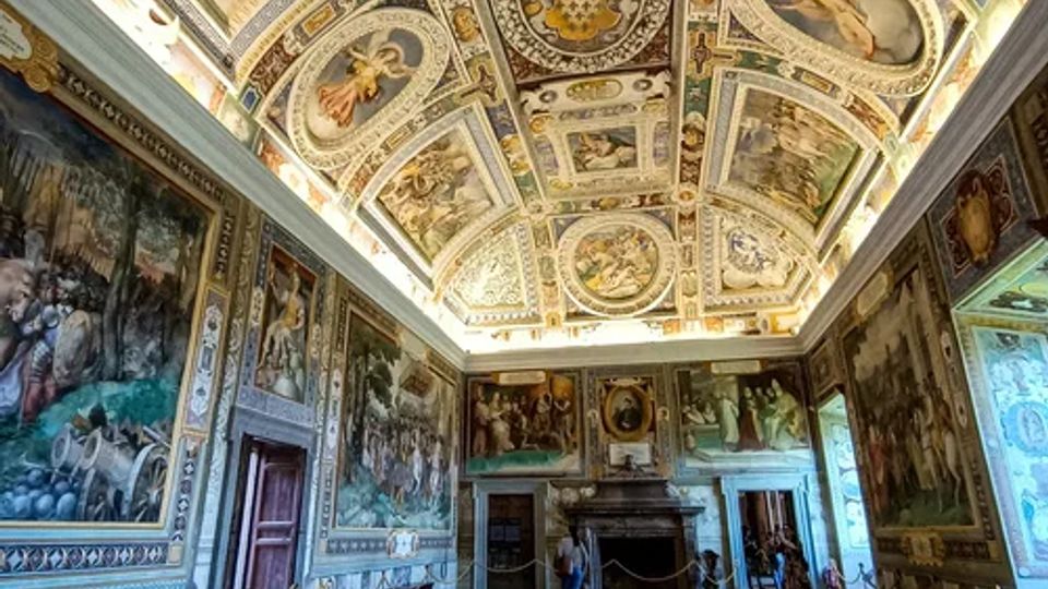 Interno di una grande sala storica con pavimento in pietra, pareti rivestite da ampi affreschi e un grande camino sul fondo. Il soffitto a volta è riccamente decorato con riquadri dipinti, motivi ornamentali e scene figurative.
