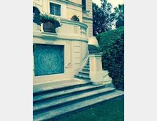 Scalinata in pietra che conduce a una grande porta decorata su una facciata chiara con balaustra e colonne, in un edificio residenziale. Ai lati ci sono siepi e alberi, con un prato verde in primo piano.