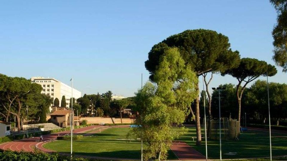Complesso sportivo all’aperto con pista di atletica rossa che circonda un campo in erba, recintato e illuminato da alti pali. Intorno si vedono alberi ad alto fusto e, sullo sfondo, un grande edificio bianco sotto un cielo sereno.