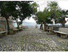 Passeggiata in ciottolato in un piccolo belvedere alberato, con panchine in pietra ai lati e ringhiera metallica sul bordo. Sullo sfondo si intravede un panorama collinare oltre un muretto in pietra.