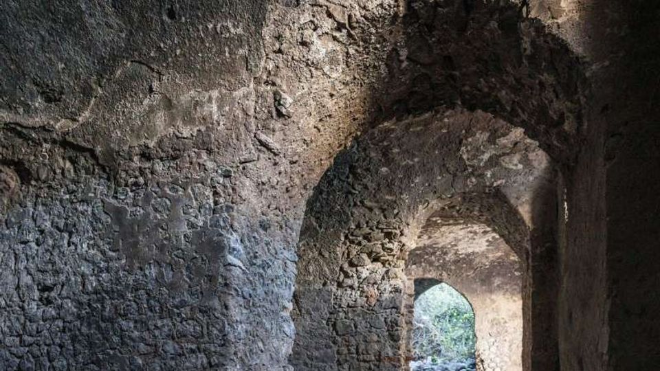 Interno di un corridoio in pietra grezza con volte ad arco e pareti irregolari. In fondo si apre un arco che conduce verso l’esterno, dove si intravede vegetazione e luce naturale.