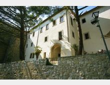 Edificio storico intonacato di bianco con più piani, finestre rettangolari e ingresso ad arco in pietra, visto dal basso dietro un muro di contenimento in pietra. L’area esterna è ombreggiata da alti pini e comprende un lampione e alcune piante vicino alla facciata.