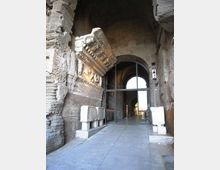 Ingresso di un antico edificio in muratura con grande arco e volte in pietra, pavimento in lastre e portale moderno in vetro e metallo. Sulla parete sinistra si vede un fregio decorativo sporgente e blocchi di pietra esposti lungo il corridoio.