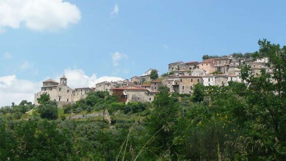 Vista panoramica di un borgo collinare con case in pietra e tetti in laterizio, disposto in salita lungo il pendio. In primo piano si vede vegetazione verde e alberi, con cielo azzurro parzialmente nuvoloso sullo sfondo.