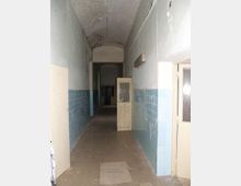Corridoio interno lungo e stretto con soffitto a volta, pareti intonacate chiare e rivestimento di piastrelle azzurre nella parte bassa. Diverse porte laterali, una porta in legno con griglia semiaperta e pavimento in cemento con segni di usura.
